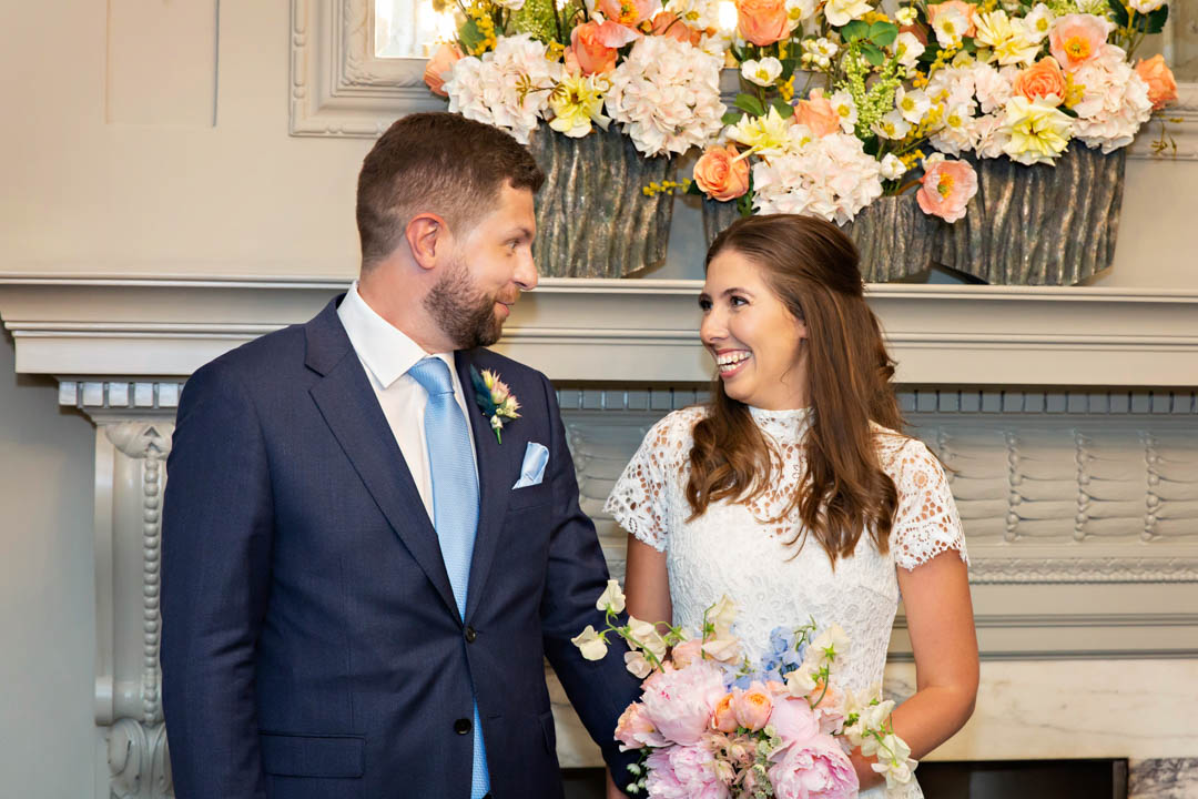 A groom and bride giggle at each other during their civil marriage ceremony in the Soho Room. The groom is wearing a blue suit with a baby blue tie and the bride is wearing a short-sleeved, high necked lace dress and holding a spring bouquet with sweet peas and peonies.