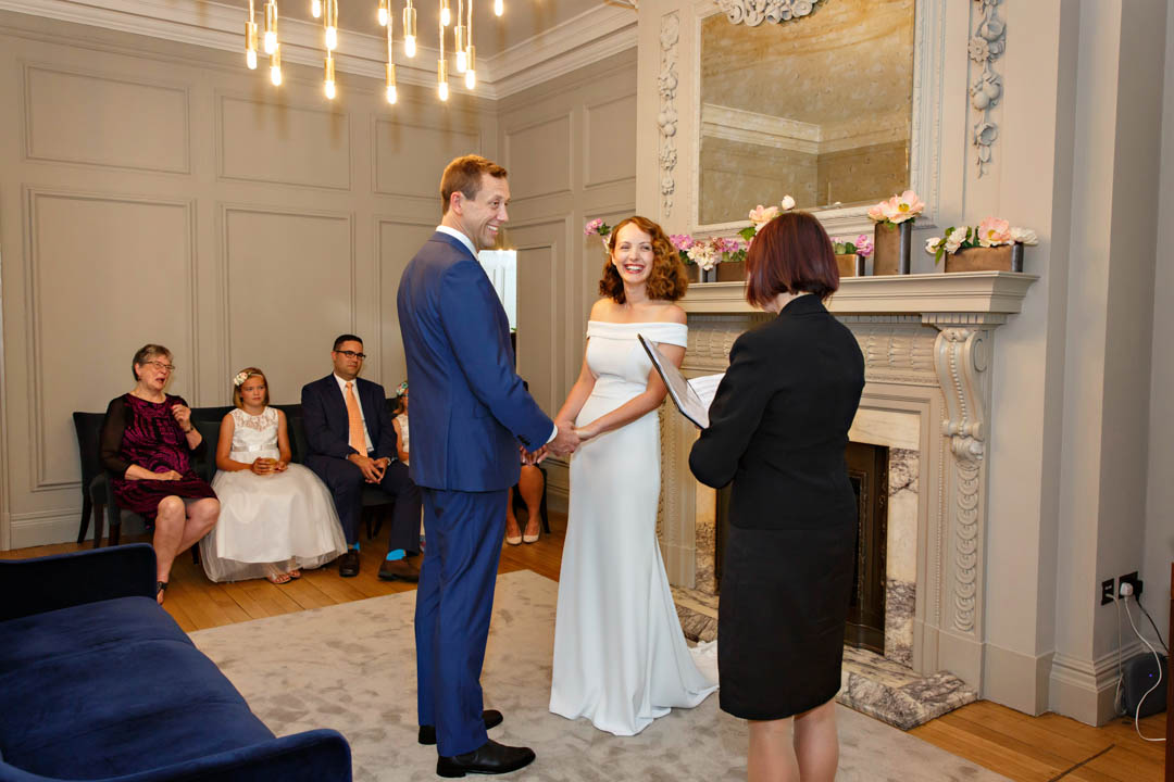 An auburn bride in an off-the-shoulder full-length dress and a groom in a navy suit, black shoes and white shirt hold hands during their wedding in the Soho Room. Guests sit in the background and the registrar, dressed in a black skirt suit is in the foreground.