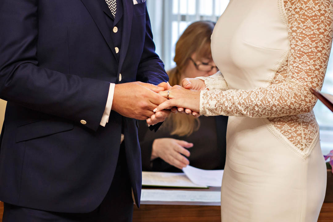 A bride and groom hold hands during the exchange of rings in the Soho Room. A registrar sits in the background.