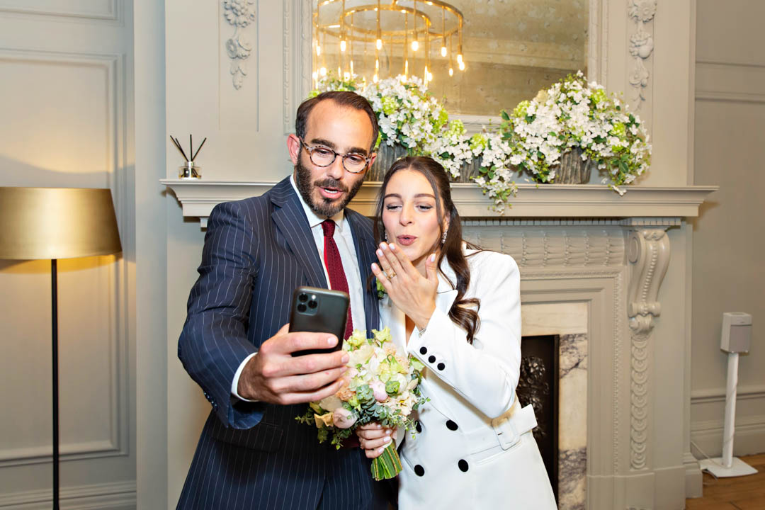 A bride wearing a white, double-breasted jacket dress and her groom in a blue pin-striped suit and dark red tie blow kisses to family on a phone video call during their small wedding in the Soho Room at Old Marylebone Town Hall.