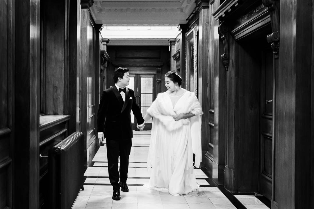 A bride wearing full-length white and a white fur stole holds hands with her groom while she admires his smart suit. They're walking along a hallway near Westminster's Soho Room, where they were married.