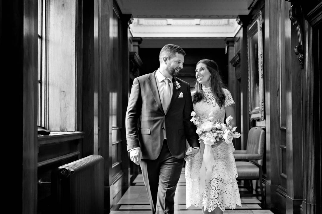 A black and white photo of a bride and groom walking along a hallway near the Soho Room. The bride is wearing a white, knee-length lace dress and carrying an avant-garde bouquet and the groom is in a suit, looking towards her.
