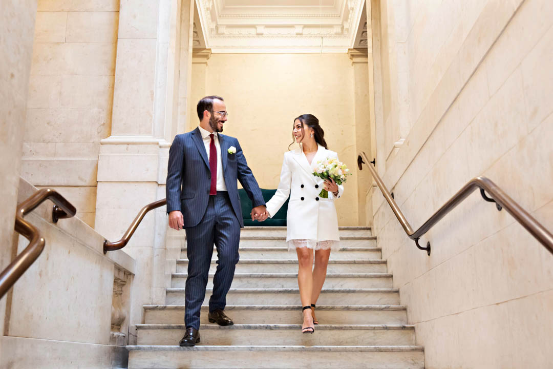 A bride wearing a white, double-breasted jacket dress walks down a marble staircase inside Old Marylebone Town Hall. She's holding the hand of her groom, who is wearing a blue pin-striped suit with a dark red tie. They're both smiling at each other after their civil marriage ceremony in the Soho Room.