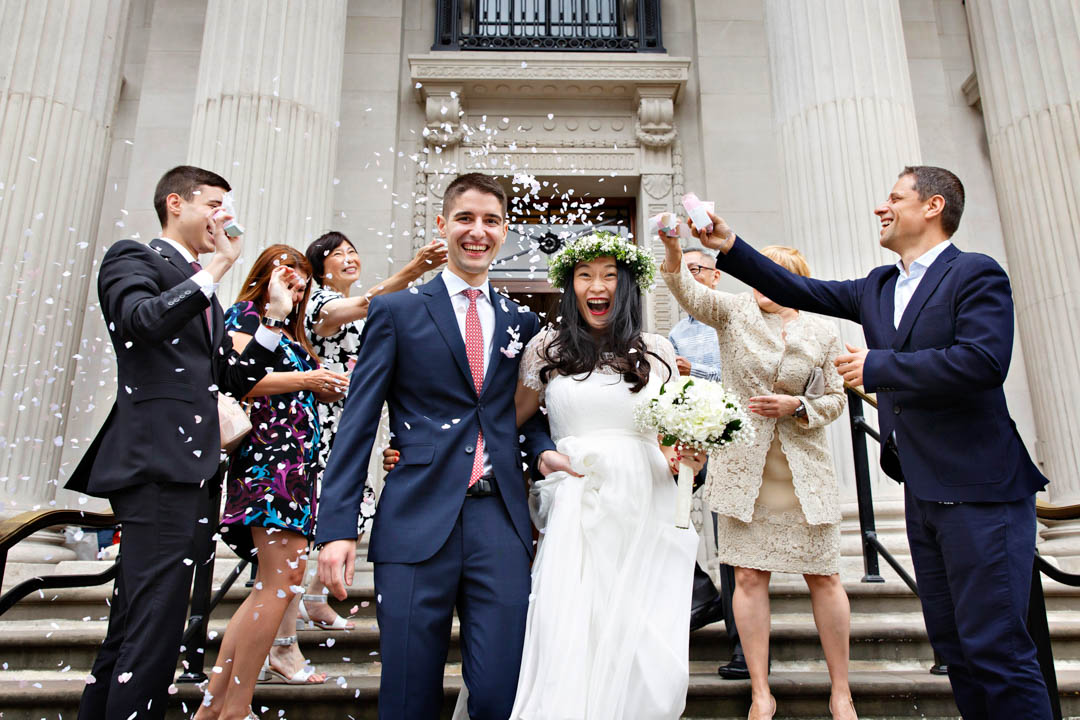 Guests throw pastel confetti over two newlyweds walking down the steps of Old Marylebone Town Hall. The bride is wearing a full-length, flowing white dress, a white fairy crown and white bouquet tied with a white ribbon. The groom has a blue suit and red tie.