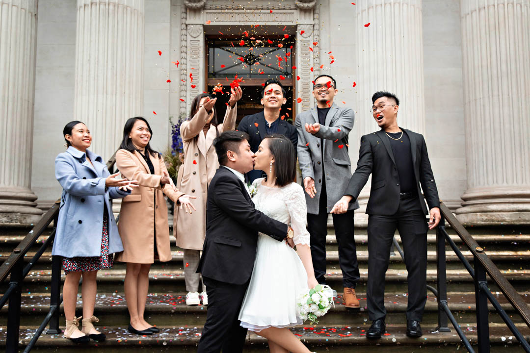 Guests throw red rose petal confetti over two newlyweds on the steps of Old Marylebone Town Hall. The bride is dressed in a knee-length white lace dress and carrying a white and green bouquet. The groom is wearing a black, three-piece suit. The bride and groom are kissing and the groom has his hands around the bride's waist.