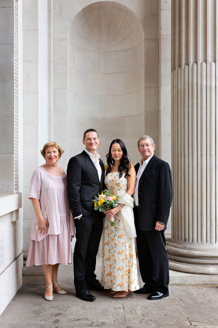 A bride in a cream dress sprigged with yellow flowers holds a yellow and pink hand-tied bouquet. Beside her is a groom in a dark suit and a bright yellow buttonhole and two family members in a traditional wedding family photo. They're standing outside Westminster's Old Marylebone Town Hall after their marriage in the Soho Room.