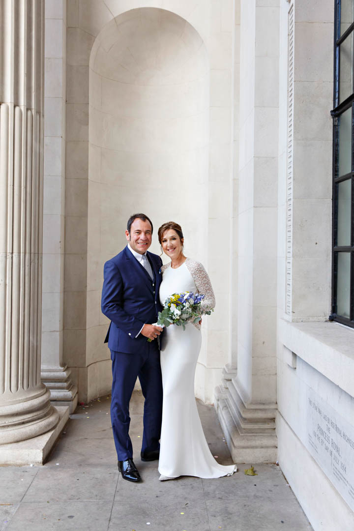 A bride in a full-length white dress with small train holds a white, mauve and yellow bouquet tied with a white ribbon. Her groom stands beside her and they hold hands. He's wearing a blue suit and matching tie. They're standing on the portico of Old Marylebone Town Hall after their civil wedding in the Soho Room.