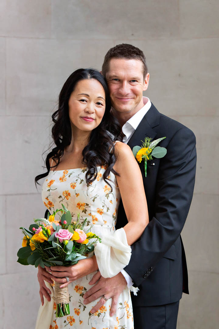 A bride in a cream dress sprigged with yellow flowers holds a yellow and pink hand-tied bouquet. Behind her is her groom in a dark suit and a bright yellow buttonhole. They're standing outside Westminster's register office after their marriage in the Soho Room.