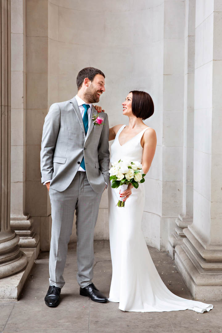 A bride in a sleeveless, v-neck full-length white bridal gown and holding a white rose bouquet leans her hand on the shoulder of her groom. They are both looking at each other and smiling. The groom is wearing a grey suit with a pink rose buttonhole and teal tie. They're standing in the portico of Old Marylebone Town Hall after their Soho Room wedding.
