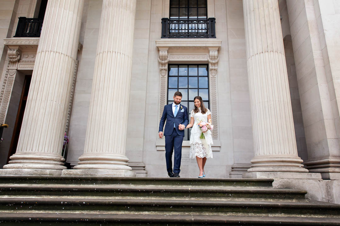 A bride in a knee-length white lace dress with short sleeves carries a pink hand-tied bouquet as she holds hands with her new husband. He is wearing a blue suit and baby blue tie. They're about to walk down the steps of Old Marylebone Town Hall after their civil marriage in the Soho Room.
