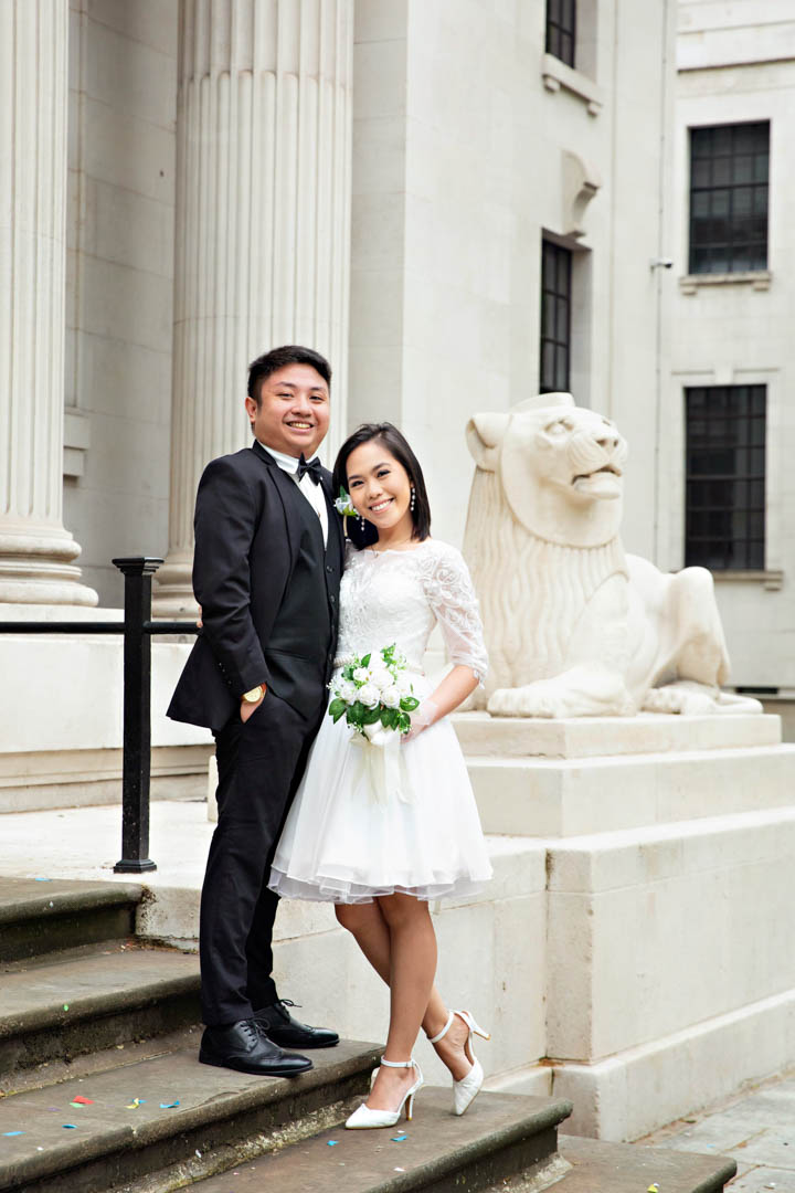 A bride in a knee-length lace wedding dress with a full skirt and white shoes stands on the steps of Old Marylebone Town Hall with her groom. He is wearing a black three-piece suit and matching shoes. In the background is one of the art deco stone lions that 'guard' the entrance.
