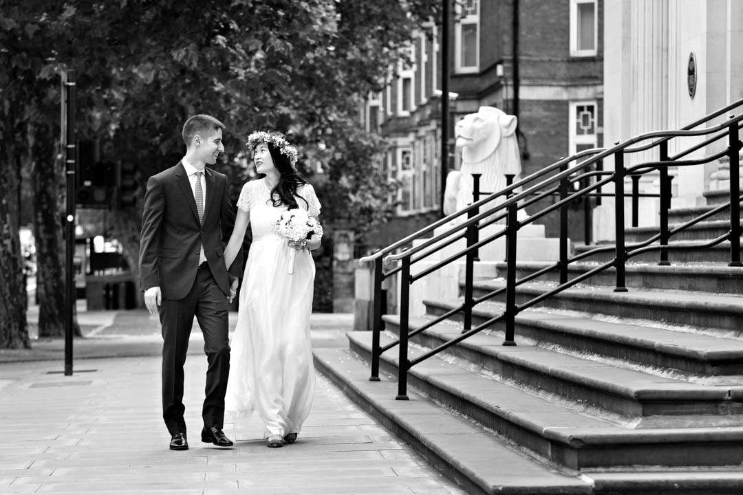 A bride in a full-length white dress and white flower crown carries a white bouquet tied with ribbon. She has her hand around her groom and he is smiling at her. They're walking along the pavement outside Old Marylebone Town Hall at 97-113 Marylebone Rd, London NW1 5PT.