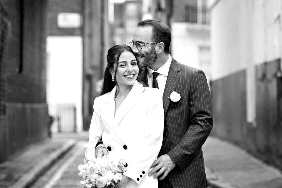 A black and white photo of a bride in a white jacket with black buttons. Her groom, in a pin-striped suit and white buttonhole, is standing behind her, with his hands on her hips and snuggling his face into her hair.