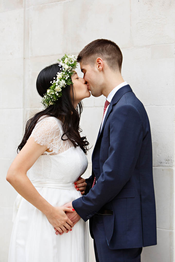 A bride in a white dress with a lace sleeve and bodice and white flower grown kisses her groom in a navy suit. They're standing beside a white wall.