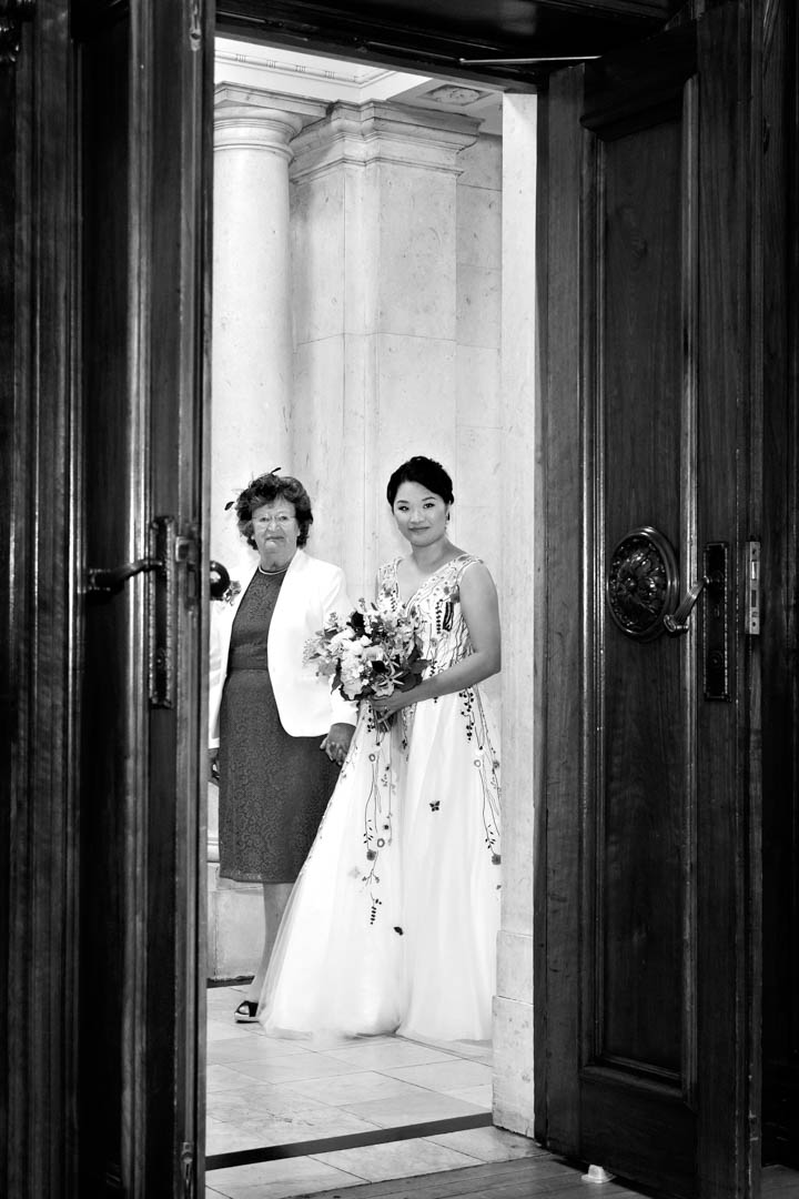 A black and white photograph through the doorframe of the Westminster Room, catching a peek at the bride and the groom's mother as the two are about to walk down the aisle together.