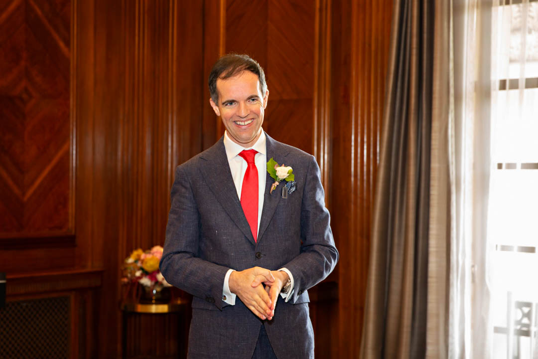 A groom smiles broadly as he watches his bride walk down the aisle towards him in the Westminster room. The bride is out of shot.