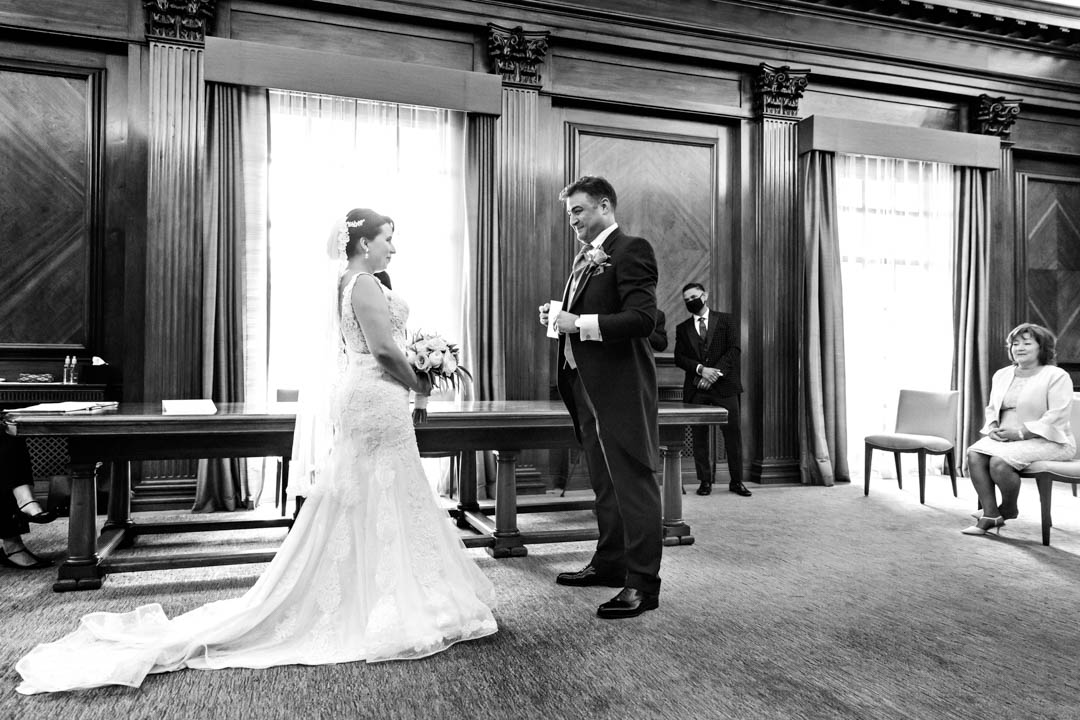 A wide angle black and white photo taken in the Westminster Room at The Old Marylebone Town Hall. The bride and groom stand facing each other as the groom prepares to read his personalised vows to the bride. The bride's full train is spread along the floor. Sunlight floods the background through two large windows in an otherwise wood-panelled room.