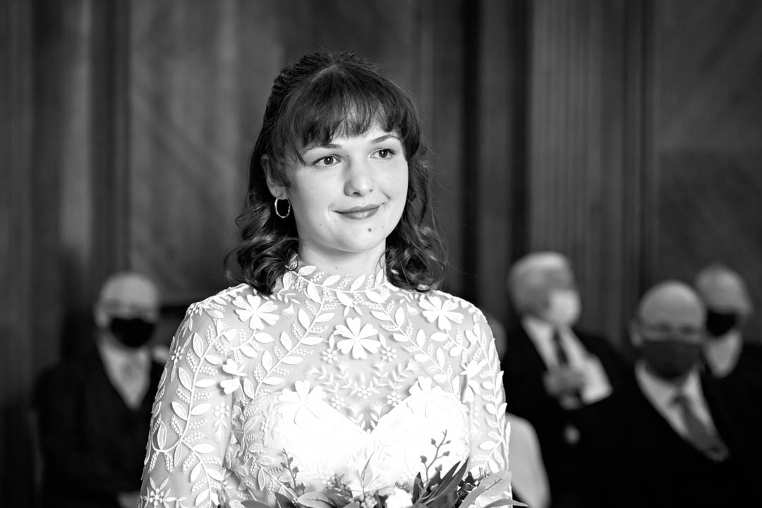 Black and white photo of a bride looking at the registrar (out of shot) as she instructs her on the wedding vows. The bride's dress features a beautiful layer of floral lace, a high neck and full sleeves over a sweetheart bodice.