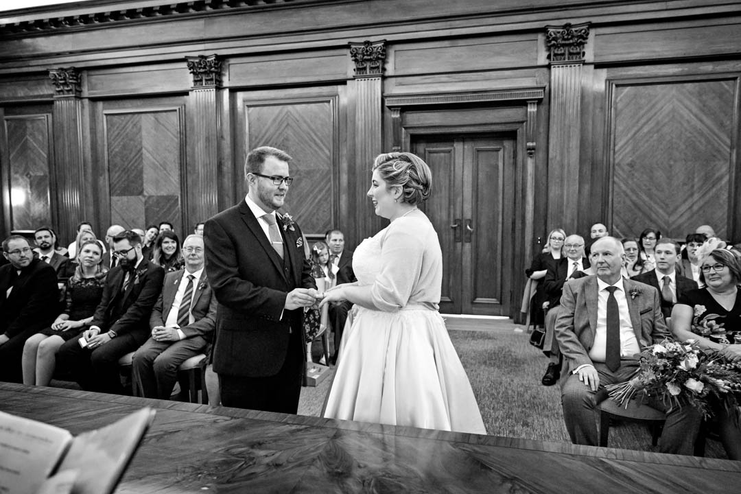 A black and white wide-angled photo within the wood panelled Westminster room. The groom is placing the wedding ring on the bride's fingers as the guests look on.