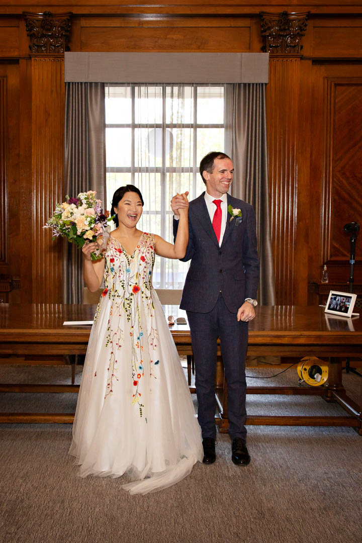 The newlyweds cheer to celebrate the end of their intimate wedding in the Westminster Room. The bride is wearing a full length dress with colourful floral detail.