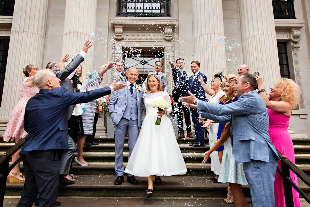 A bride and groom are surrounded by their guests throwing confetti on the steps of Old Marylebone Town Hall. They just got married in the Westminster Room.