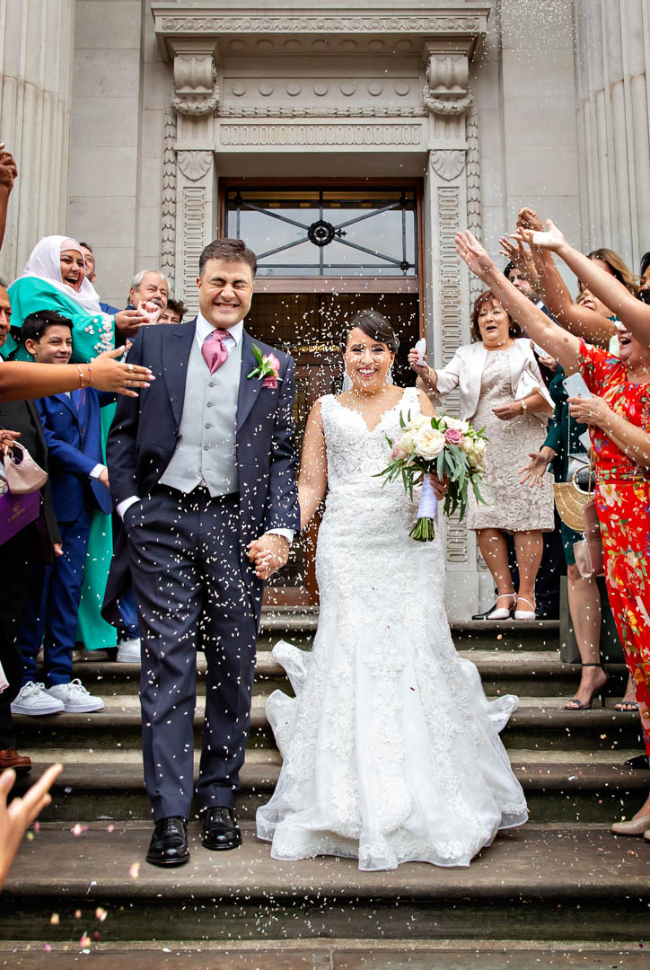 A full length image of a bride and groom exiting their Westminster room wedding into lots of confetti thrown by their guests.