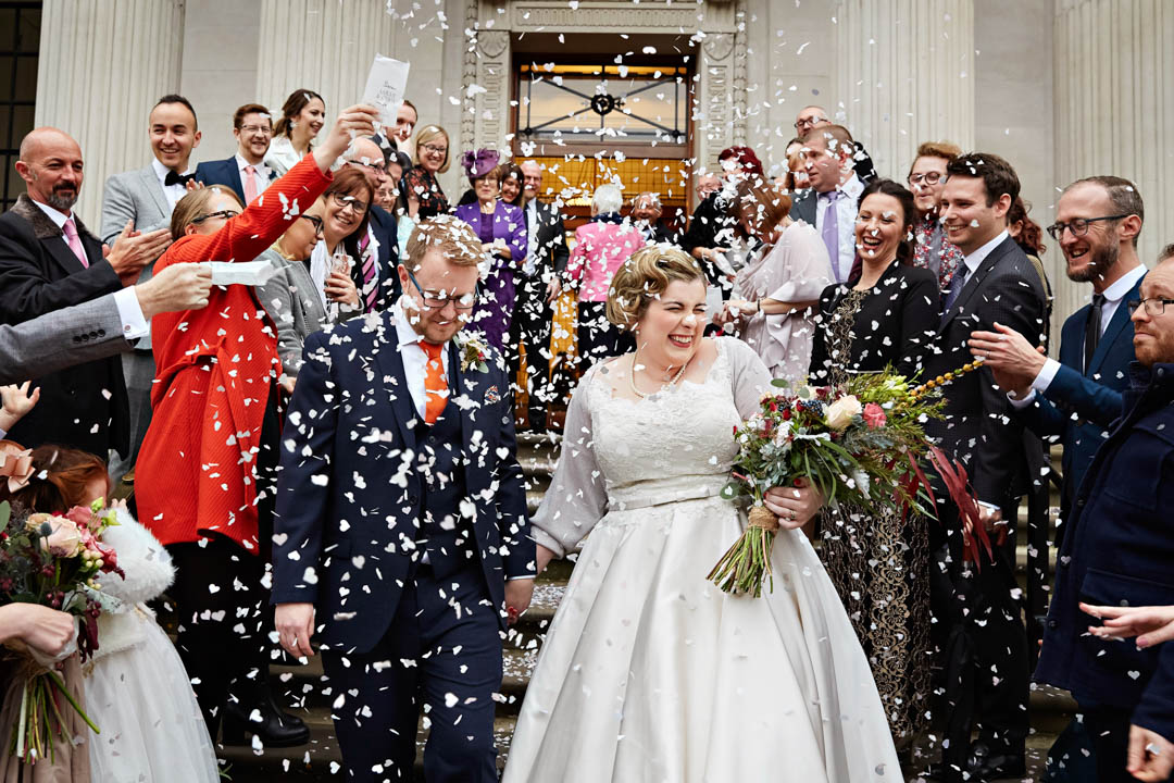 A bride in a 1950s style vintage dress and hairstyle walks out into a flurry of confetti, holding her groom's hand. They just got married in the Westminster room.
