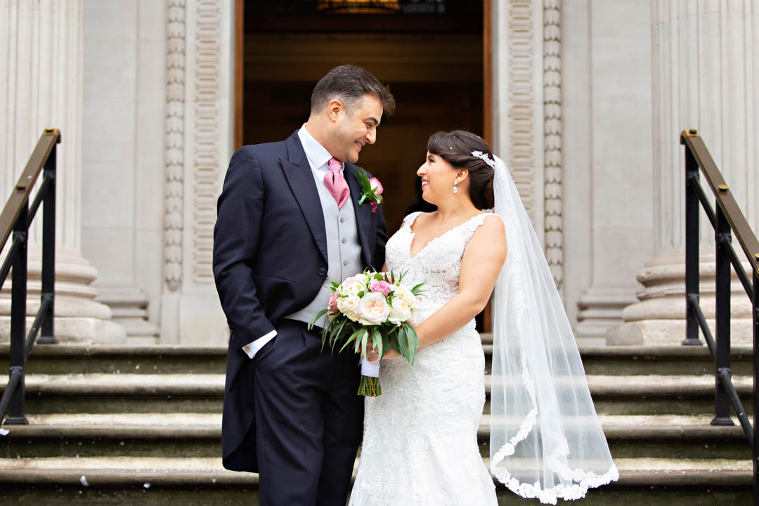 A bride and groom smile at each other on the steps of Old Marylebone Town Hall, as they exist from their Westminster Room wedding celebration.