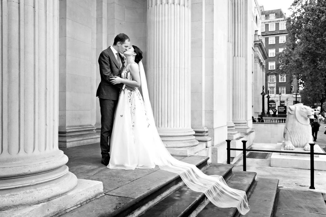 A wide black and white photographs of a groom kissing his bride at the top of the stairs of The Old Marylebone Town Hall. The brides vail flows down the stairs.