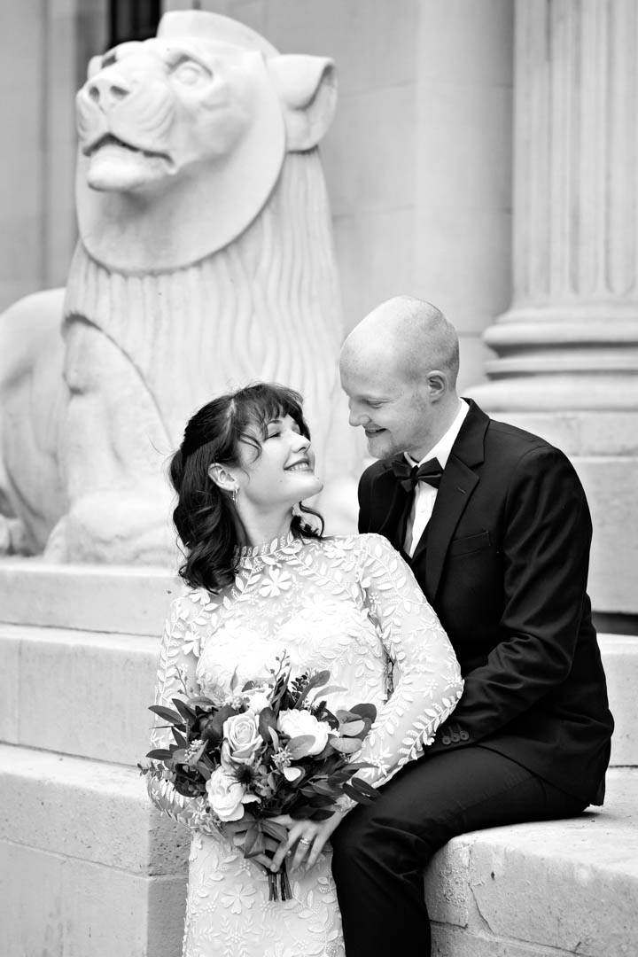 A black and white photograph of a bride and groom cuddling in front of one of the stone lions at The Old Marylebone Town Hall after getting married in the Westminster room. The bride is wearing a fitted, long sleeved lacy wedding dress, and she's looking back over her shoulder at the groom.