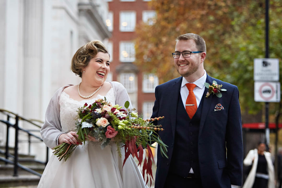A bride and groom laugh at each other as they walk along the pavement in front of The Old Marylebone Town Hall. The bride is carrying a large bouquet of flowers in autumnal colours of burgundy, peach and green. Her hair is set in a 1950s vintage style. The groom is wearing a navy tweed 3 piece suit.