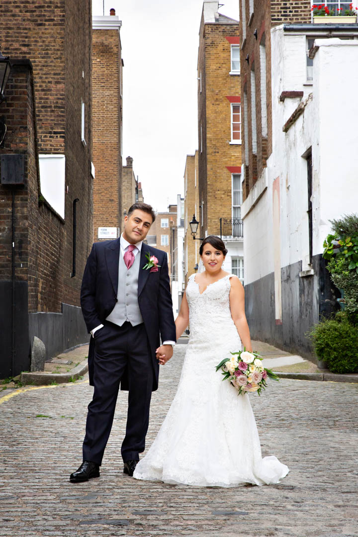 A bride stands hand in hand with her newly wed groom. The bride is wearing a sleeveless wedding dress whilst the groom is in a black morning coat with grey waistcoat and pink tie.