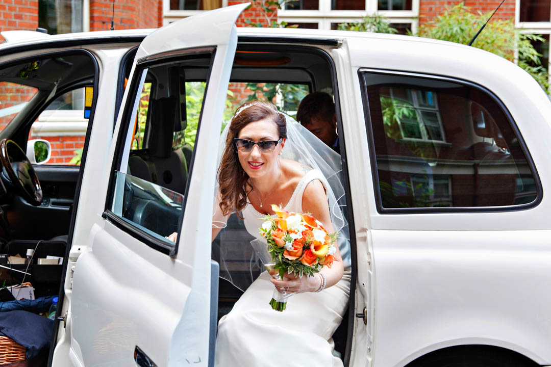 A super fashionable bride wearing cats eyes sunglasses arrives at Mayfair Library in a white wedding taxi. She's getting out of the taxi carrying a burnt orange and white bouquet.