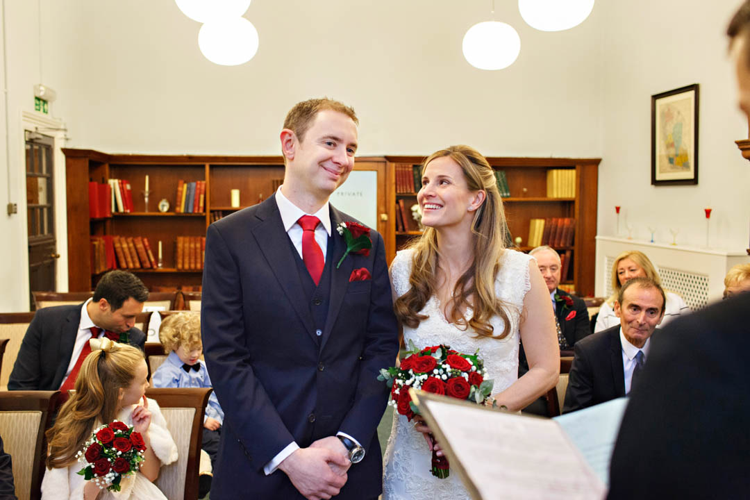 A bride looks loving at her groom as he confirms his name for the registrar at the start of their intimate wedding ceremony in the Marylebone Room at Mayfair Library.