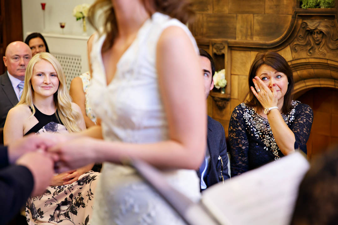 A bride's mother wipes away a tear of joy as she watches her daughter get married to the man she loves. The bride's body can be seen out of focus in the foreground, with the mother of the bride clearly visible in the background.