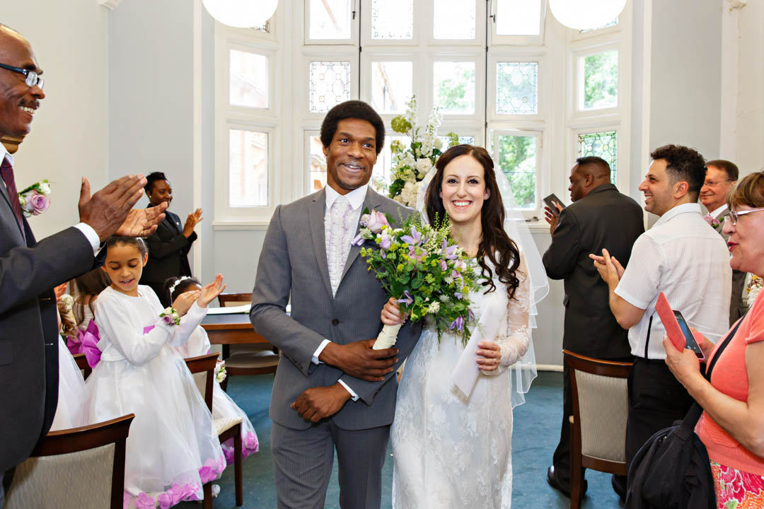 A beaming bride and groom exit their register office wedding ceremony as husband and wife, with their guests standing up and applauding.