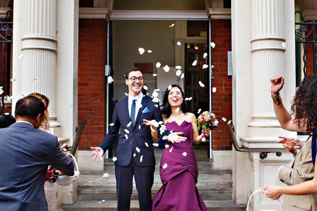 A bride wearing a purple strapless gown and her groom are surprised by their guests flowing confetti made from large white rose petals.