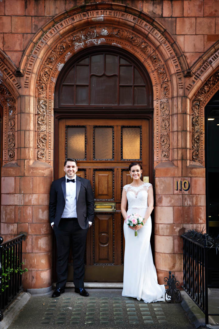 An elegant bride and groom use the classic redbrick Mayfair architecture as a backdrop for a stylish wedding portrait. The groom is wearing a black tuxedo and the bride is wearing a fitted, sleeveless, lace dress and holding a pastel bouquet of roses.