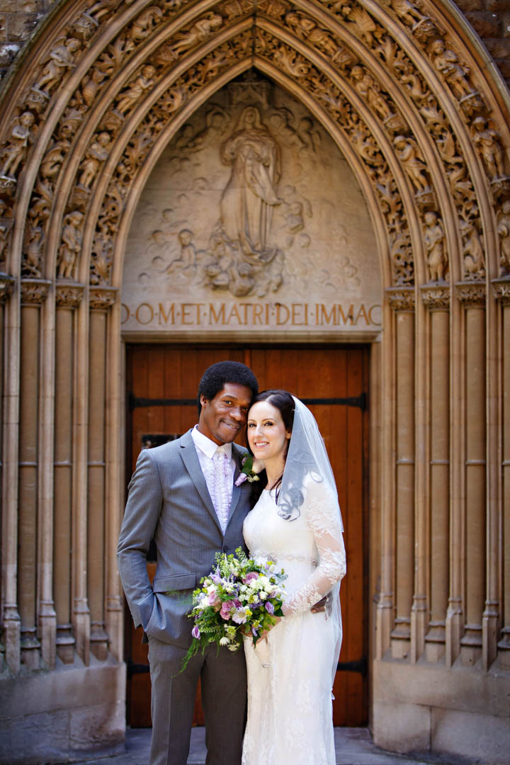 A classic, timeless wedding portrait of a bride and groom in front of the ornate stone carved entrance way to Farm Street Church. The groom is wearing a vintage ruffled white shirt under a grey suit and is sporting a tightly cropped afro hair do.