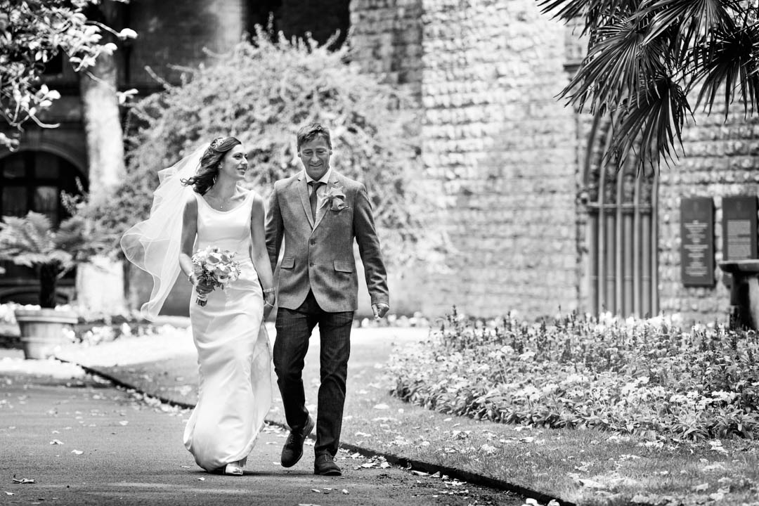 a black and white photograph of a bride and groom walking through the park hand in hand. They look unobserved and the bride's dress and veil are billowing in the breeze.