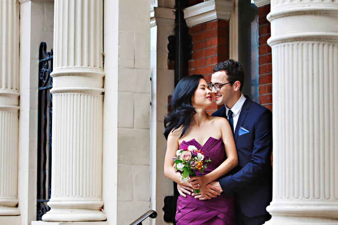 A bride share an intimate moment during their relaxed wedding couples' portrait session. They are in a tight embrace wit their lips almost, but not quite, touching for a kiss, amongst the stone pillars of Mayfair Library.