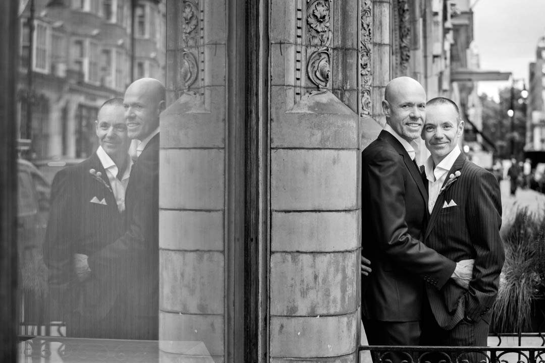 A creative and timeless black and white photograph of two gay grooms embracing each other while their complete reflection appears in the shop window adjacent to the two.