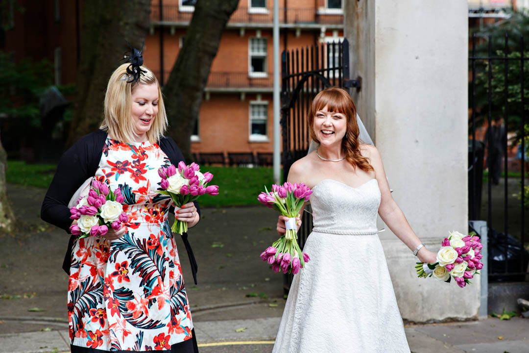A bride walks towards Mayfair Library with her bridesmaid, carrying not only her own bouquet, but also all the bridesmaids bouquets. The flowers are white roses and pink tulips.
