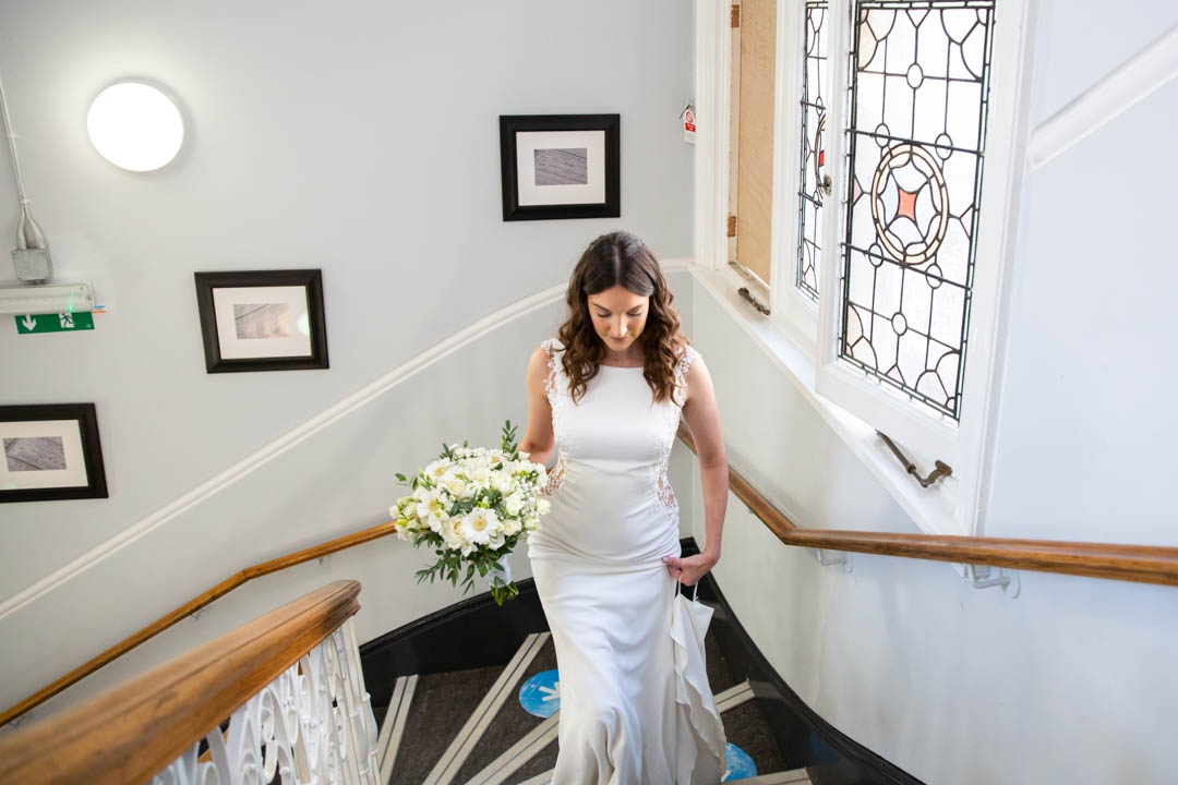 A wide-angle image of an elegant bride in a pronovias dress walking up the stairs of Mayfair Library on her own.