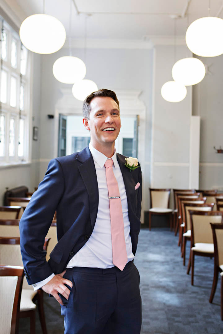 A groom stands smiling in the aisle of the Mayfair Room at Mayfair Library, having just finished his private pre-ceremony interview with the Westminster registrars. He's now ready to get married.