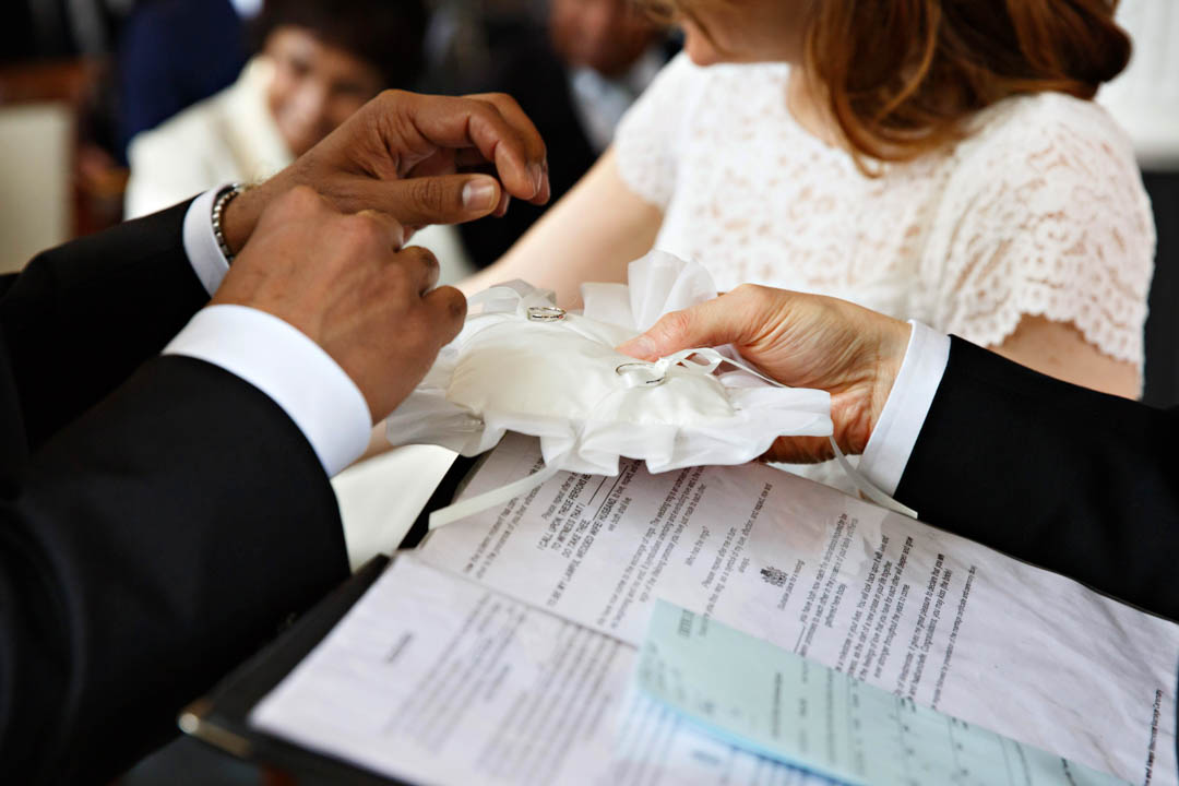 A closeup photograph of the registrar presenting the wedding rings to the groom on a special wedding ring pillow. Both rings are clearly visible as the groom reaches for the bride's ring.