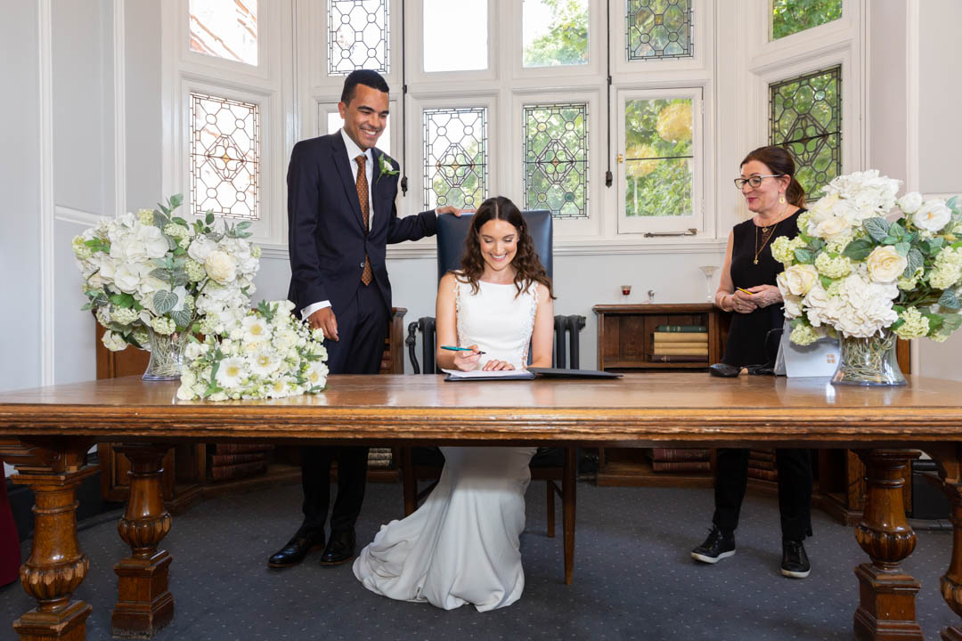 A bride is seated at a very wide table, getting ready to sign the wedding register book. The groom and the Westminster registrar pay close attention.