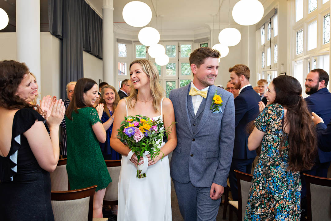A bride and groom smile at their wedding guests as the exit up the aisle of the Mayfair Room at Mayfair Library among a big round of applause from all their wedding guests.