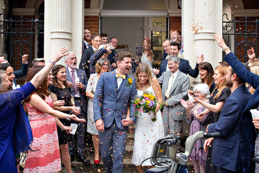 Newlyweds are surrounded by their guests throwing confetti on the steps of Mayfair Library.
