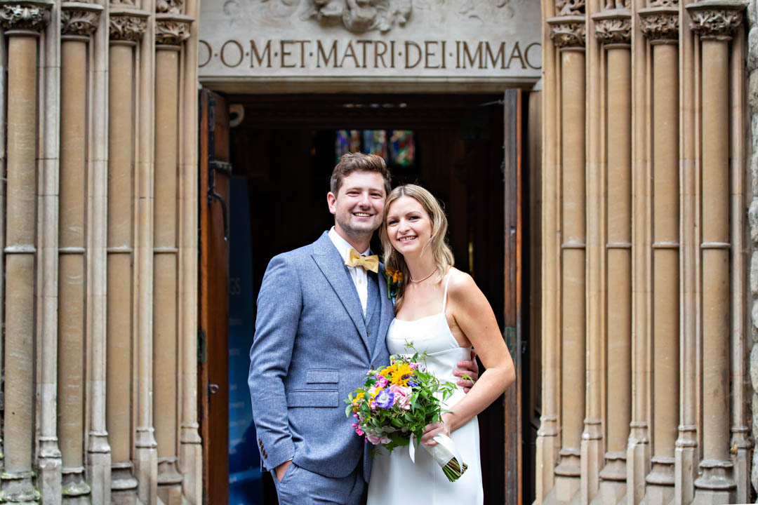 A bride and groom pose for a wedding portrait framed by the ornate fluted stone carved entrance to Farm Street Church in Mayfair.